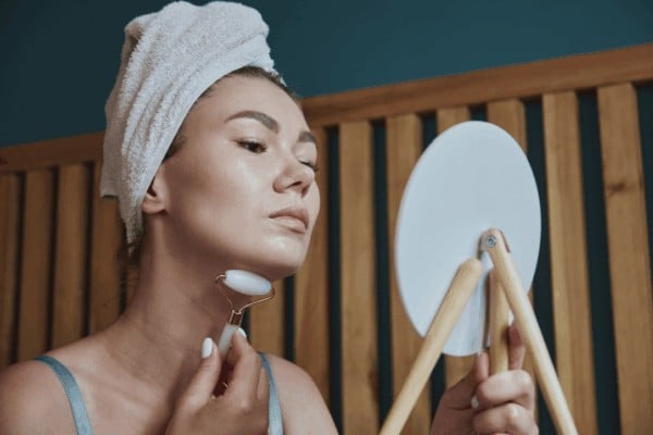 Woman with a hair towel using a crystal beauty tool on her jawline while looking in a mirror during a nighttime skincare ritual.